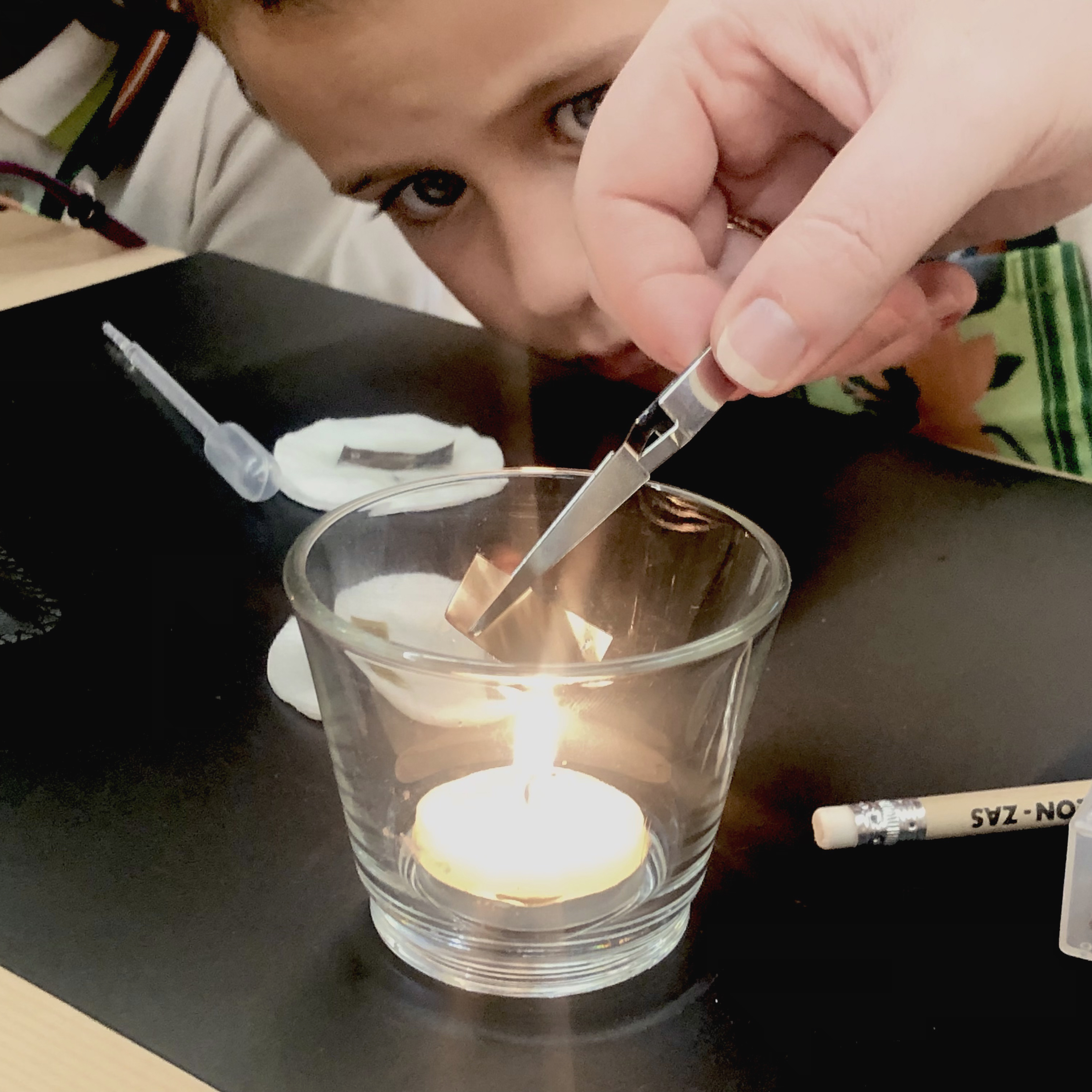 Detalle de una mano exponiendo una muestra de metal al calor de una vela. Un niño lo observa con atención.